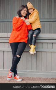 Vertical shot of pretty beautiful young woman dressed in red sweater and ragged jeans, stands closely to her small daughter who embraces her, enjoy togetherness. Parenthood and childhood concept