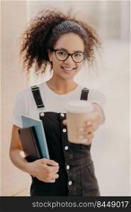 Vertical shot of pretty female with curlly hair, holds takeaway coffee, has drink during break at university, wears glasses and denim overalls, expresses positive emotions. Happy student with beverage