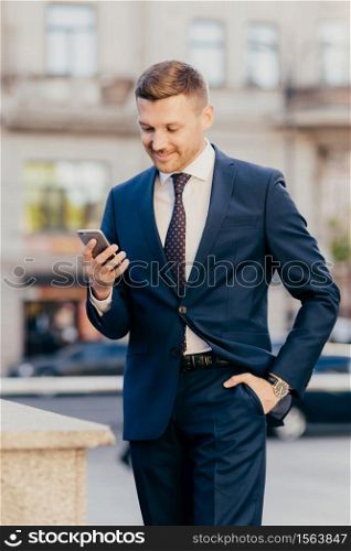 Vertical shot of prosperous male manager wears elegant suit, keeps hand in pocket, uses smart phone, being always in touch, reads business news on internet website, concentrated into screen.