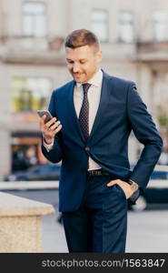 Vertical shot of prosperous male manager wears elegant suit, keeps hand in pocket, uses smart phone, being always in touch, reads business news on internet website, concentrated into screen.