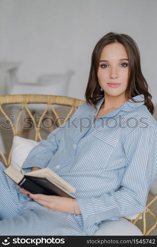 Vertical shot of relaxed beautiful woman dressed in domestic clothes, holds book in hands, enjoys reading in calm atmosphere, looks with calm atmosphere at camera. People, rest, lifestyle concept