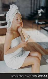 Vertical shot of relaxed happy woman applies moisturising cream on face, cares about complexion and skin, sits over domestic atmosphere, wrapped in bath towel. Facial treatment and hygiene concept