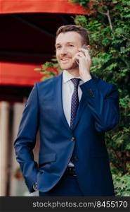 Vertical shot of satisfied happy male employee has telephone conversation, dressed in black suit, keeps hand in pocket, has glad expression, gentle smile on face, being confident in everything