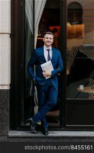Vertical shot of successful male executive worker in formal suit, keeps hand in pocket, keeps legs crossed, holds journal, stands near cafe, has positive expression. Business and work concept