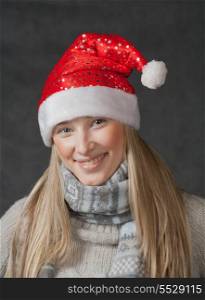 Vertical shot of the smiling blond haired 20s women weared Santa hat with stars pattern. Cute blond wearing christmas hat on dark background