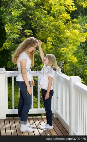 Vertical side view of younger sister measuring height difference with older sister while outdoors on patio with blurred out trees in background