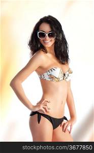 very beautiful sensual woman with long black hair sunglasses and stone necklace in fashion swimsuit, she is turned of three quarters at left, looks in to the lens and smiles and her right hand is on the right hip