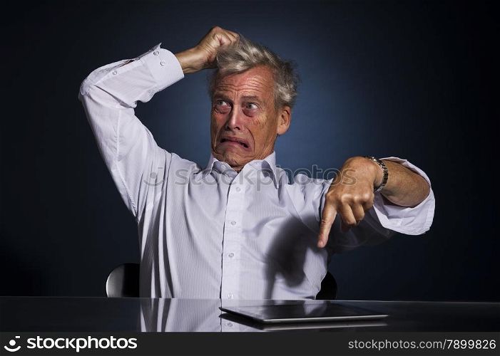 Very emotional upset frustrated senior business man pointing at his tablet computer lying on the desk while tearing at his hair with his hand