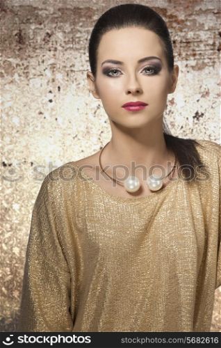 very pretty female with long brown hair wearing elegant golden dress, posing with stylish make-up and big trendy necklace.