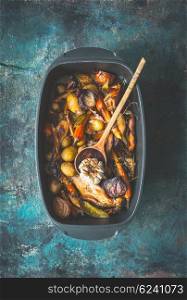 Very rustic roasted vegetables stew or ragout with wild game and wild fowl and forest mushrooms in black backing dish with wooden cooking spoon on dark aged background, top view. Country food