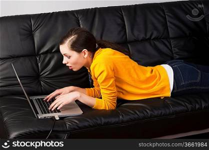 very young girl in yellow shirt laying an a black sofa and working on laptop computer, she&acute;s liyng on the the sofa, watches the laptop&acute;s display with expression of surprise