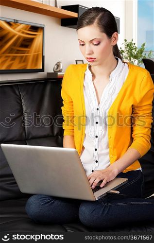 very young girl in yellow shirt laying an a black sofa and working on laptop computer, she&acute;s on her knees on the sofa and watches the laptop&acute;s display