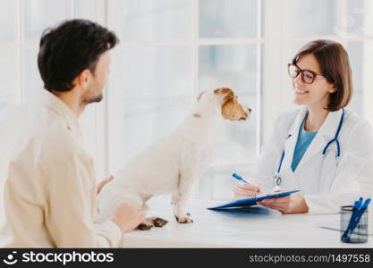 Vet checks pet in hospital, modern office, communicates with male host, writes down form in clipboard, likes animals, cares about health. Dog owner in doctors cabinet with jack russell terrier