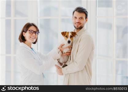 Vet female and male pet owner poses with favourite dog, come to veterinary office or hospital for doctor checkup, stand together indoor against big window, talk to each other, give useful advice