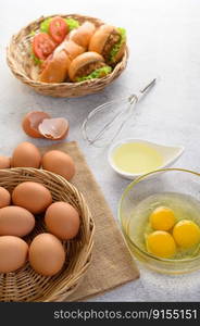 Vettical image and Selective focus yolk in glass bowl and in wicker basket, blurred hamburgers and hotdog with slice tomato and lettuce in weave basket, oil in cup and egg whisk placed nearly