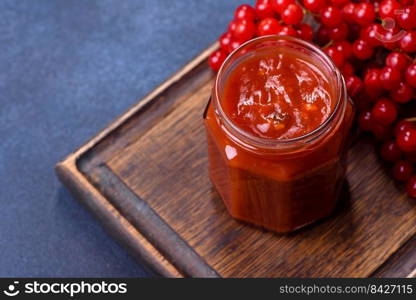 Viburnum fruit jam in a glass jar on a dark concrete background near the ripe red viburnum berries. Source of natural vitamins. Viburnum fruit jam in a glass jar on a dark concrete background