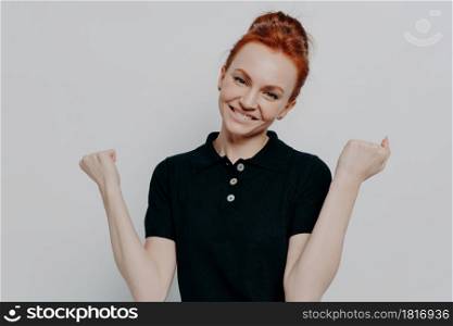 Victory concept. Studio shot of happy caucasian redhead woman clenching fists and smiling happily, excited ginger female in black casual t shirt achieving goal, isolated over grey background. Studio shot of happy caucasian redhead woman clenching fists and smiling happily