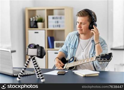 video blogging, music and people concept - young man or musician in headphones with camera videoblogging and playing guitar sitting at table at home. man or blogger with camera playing guitar at home