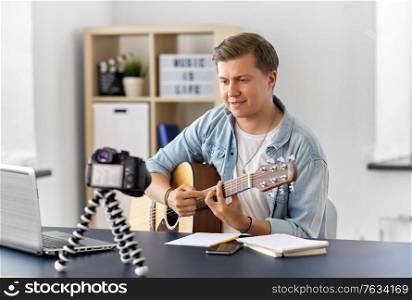 video blogging, music and people concept - young man or musician with camera videoblogging and playing guitar sitting at table at home. man or blogger with camera playing guitar at home