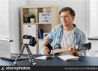 video blogging, music and people concept - young man or musician with camera videoblogging and playing guitar sitting at table at home. man or blogger with camera playing guitar at home