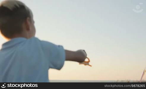 A boy launches a kite in a strong wind. In the background, a kite flies in the sky. Sunset.