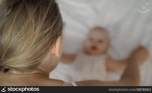 A fair-haired woman doing some playful exercises with her six-month-old baby daughter who is lying in front of her on white bed linen. The woman is holding the baby's arms with her hands and moving them from side to side. Even though the baby is a bit blurred, it is obvious that she is having a good time.