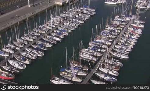 A marina; white sailboats moored at docks in a harbor side by side. A busy road directly by the harbor.