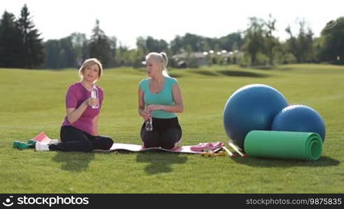 Active fitness senior women in sportswear having a break, drinking water and relaxing during training in park while sitting on exercise mat on green lawn. Sporty athletic adult females taking a break to hydrate during outdoors workout on a sunny day.