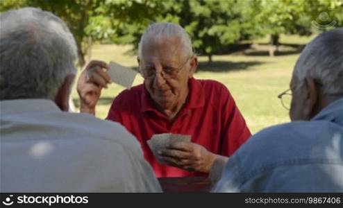 Active retired people, old friends and leisure, group of four old men having fun and playing cards game at park. Sequence of wide and medium shot