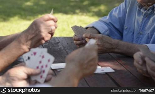 Active retirement, old people and seniors free time, group of four elderly men having fun and playing cards game at park. Sequence of closeup, medium and wide shots