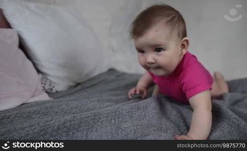 Adorable baby girl playing on bed, making her first step, learning to crawl. Portrait of happy infant child having fun on bed while family relaxing at home. Slow motion.