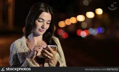 Adorable brunette female typing message on smartphone and smiling while standing on night city street against streetlights bokeh background. Cheerful stunning long hair woman in elegant clothes using mobile phone over night city lights. Shallow DOF.