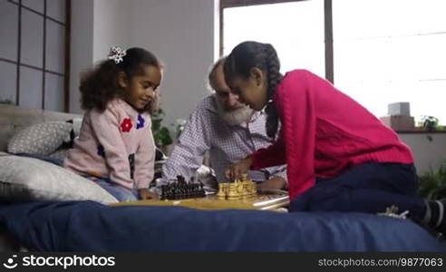 Adorable little sisters playing chess with the help of a handsome grandfather while sitting on a bed at home. The pensive girl is moving a pawn, and her younger sister is thinking intensely about the move. The caring grandpa is giving advice to his granddaughter about the game.