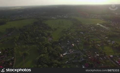 Aerial high level flight above the countryside in Russia. View of forest, country houses and agricultural fields against blue sky with clouds in sunset time at summer