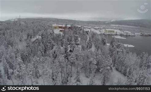 Aerial shot of winter village in pine wood by the lake in Finland. Landscape with snow covered evergreen trees, water and hills in the distance