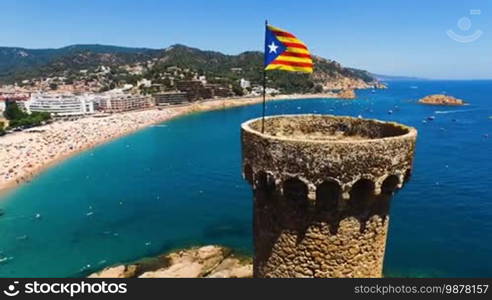 Aerial view of an old architectural building with tourists on the beach and yachts in harbor. The national flag is waving on the castle. Spain, Catalonia, Castell de Tossa