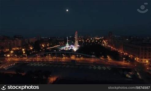 Aerial view of night illuminated Valencia with car traffic on the roads and luminous amusement park in green city area