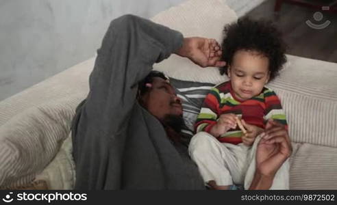 Affectionate African American father with dreadlocks caressing curly mixed race toddler son's hair while lying on the sofa. Loving dad and little child spending time together, father tenderly stroking son's hair. Slow motion.