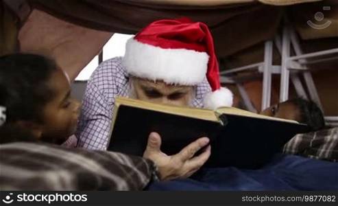 Affectionate grandfather in Santa hat reading a book to his mixed-race granddaughters as they lie down in a cubby house made of blanket and chairs. Cheerful grandpa and grandchildren reading fairy tales together on Christmas Eve. Dolly shot. Slow motion.