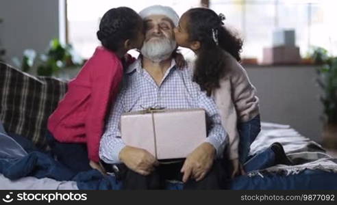 Affectionate little mixed race granddaughters kissing excited grandfather with gift on his lap on the cheeks and embracing at Christmas. Little girls giving a kiss to grandpa in Santa hat while happy family spending time together on winter holidays.