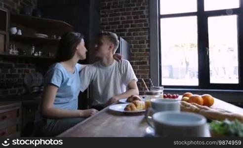 Affectionate young hipster kissing his beautiful girlfriend while sitting at the kitchen table and having breakfast in the morning. Happy attractive couple in love spending lovely morning together in modern kitchen.