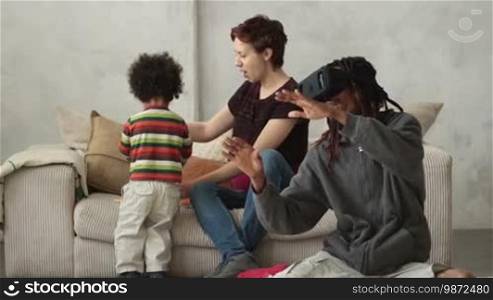 African American hipster with dreadlocks sitting on the floor and having fun with virtual reality goggles headset glasses while his Caucasian wife playing with mixed race son on background. People using new trends technology. Slow motion.