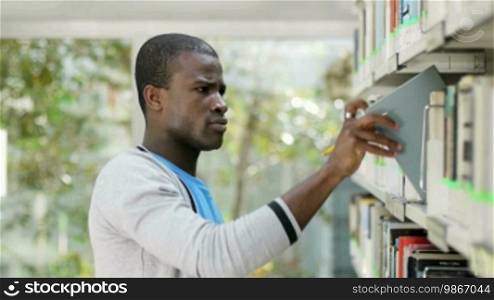 African American male college student taking book from shelf in library and looking at camera