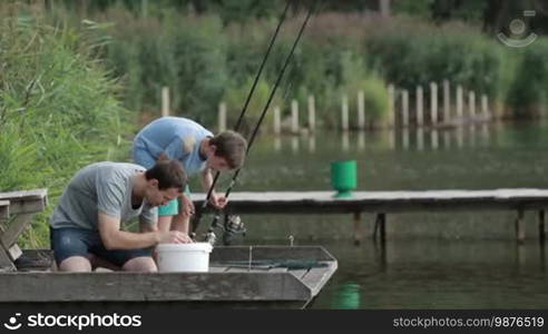 Angler kneeling on wooden pier to bait hook on fishing rod while fishing together with teenage son on the pond on sunny summer day. Father and boy spending leisure fishing against rural landscape background. Side view. Slow motion.