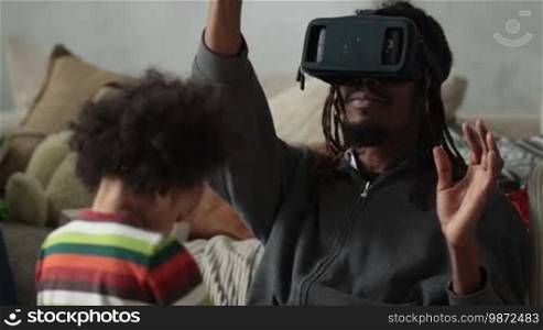 Another reality is here! Handsome young African man in VR headset sitting on the floor and gesturing in domestic interior. Hipster with dreadlocks using virtual reality goggles while mixed race son playing with toys. People using new technology.
