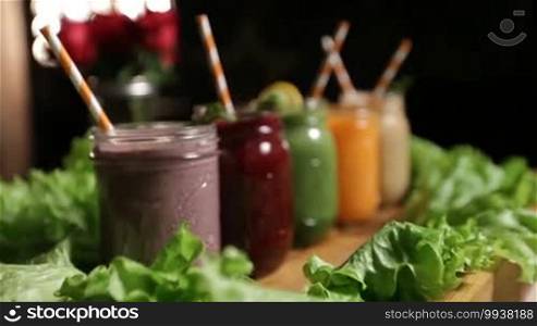 Assortment of fruit and vegetable smoothies in mason jars with striped straws, mint leaves, and lemon slices on a wooden tray decorated with a green salad circle close-up. Healthy vegetarian diet for weight loss and detox.