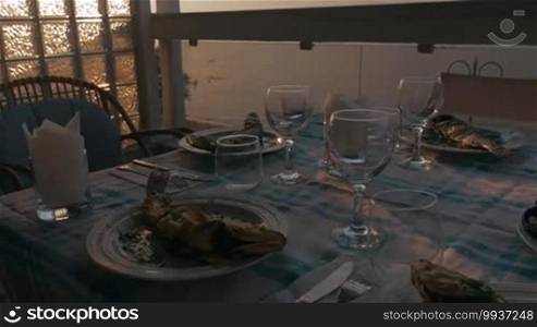 At sunset in the city of Perea, Greece, a dinner table served with cooked fish, snacks, and glasses for wine. Enjoy a beautiful sunset and the coast of the sea.
