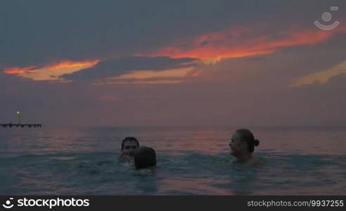 At sunset, mother, father, and their son are swimming in the sea in the city of Perea, Greece. They are splashing in the water and having fun.
