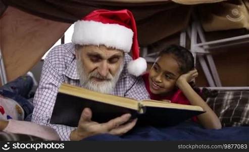 Attentive handsome grandfather with two mixed race little sisters reading fairy tales on Christmas Eve while lying together in cubby house. Caring grandpa in Santa hat and cute granddaughters reading an interesting book on winter holidays. Slo mo.