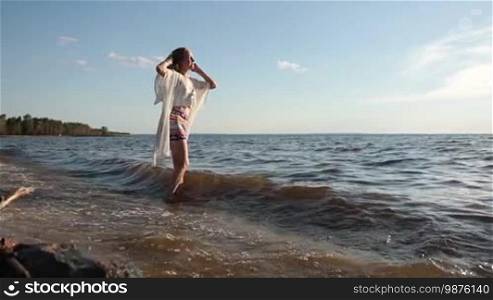 Attractive blond female in casual clothes delighted with seascape and setting sun during summer vacation on seaside. Beautiful serene relaxing woman in pure happiness and elated enjoyment with arms raised outstretched up. Slow motion. Stabilized shot